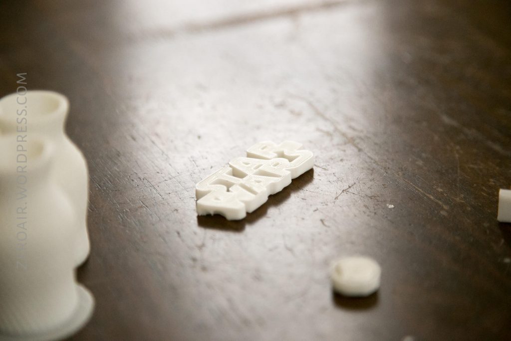 A 3D-printed object with the Star Wars logo lies on a wooden surface, surrounded by other small white plastic pieces. The text ZOKOAIR.WORDPRESS.COM is visible along the left side of the image.
