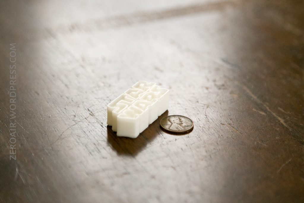 A white 3D-printed object with the Star Wars logo sits on a wooden surface next to a U.S. quarter, showing the objects size. The text ZEROAIR.WORDPRESS.COM is visible along the left edge.