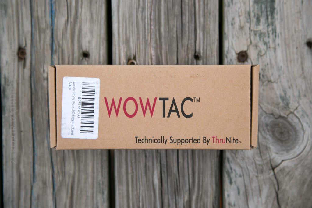 A rectangular cardboard box labeled WOWTAC Technically Supported By ThruNite sits on a weathered wooden surface. A barcode sticker is attached to the left side of the box.