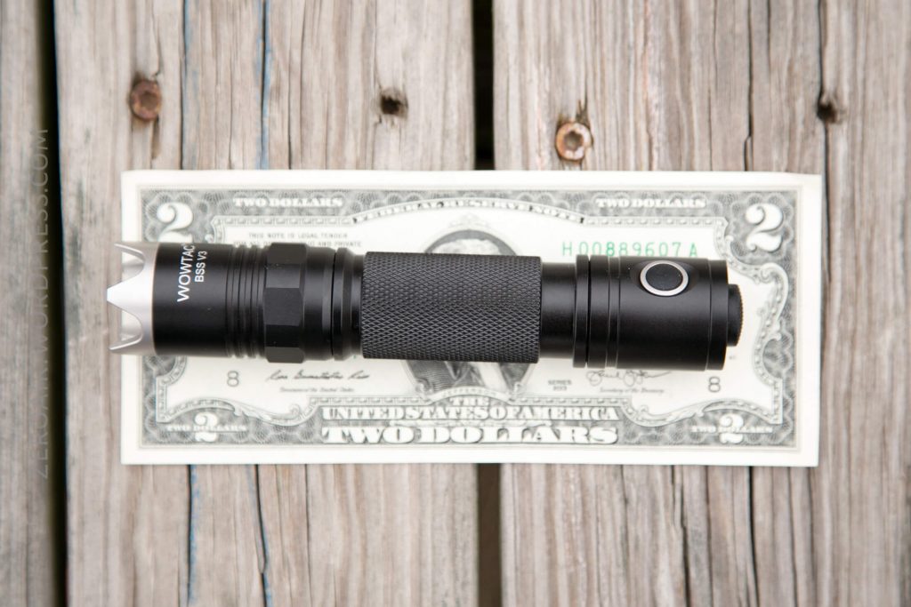 A black flashlight is placed horizontally on top of a two-dollar bill, both resting on a wooden surface with visible grain and gaps between the planks.