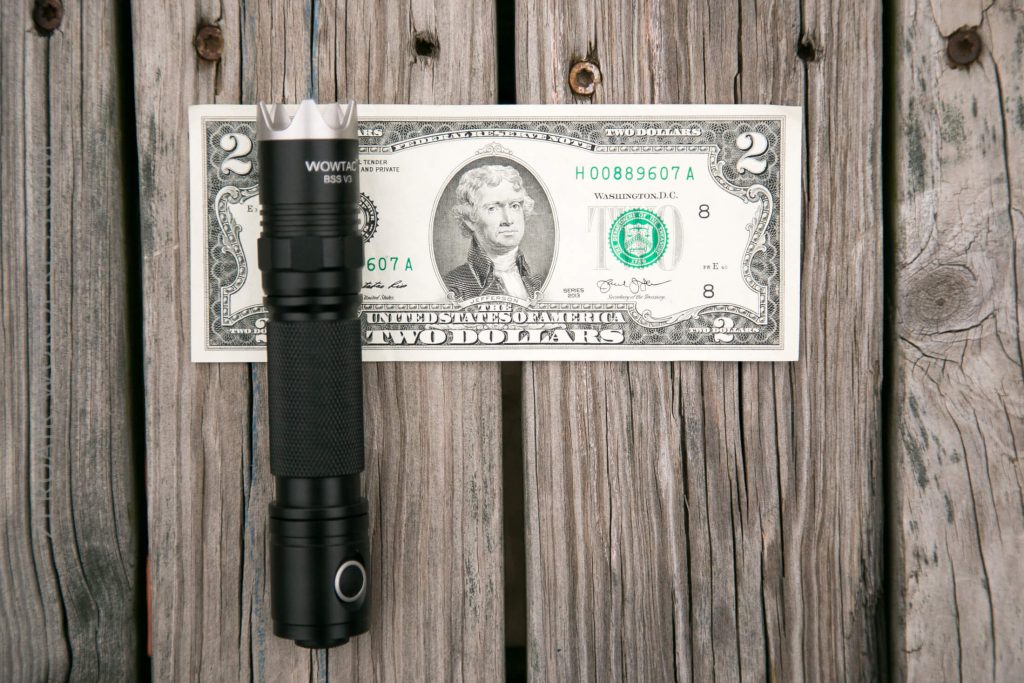 A black flashlight is placed vertically next to a two-dollar bill on a weathered wooden surface. The flashlight is slightly taller than the bill.