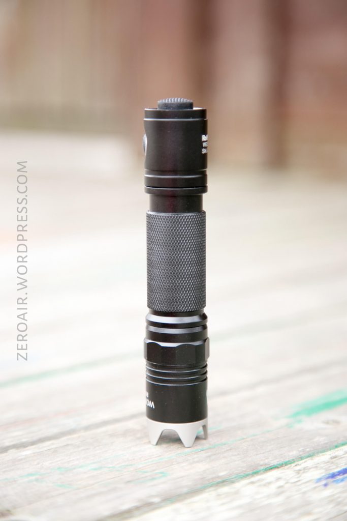 A black tactical flashlight standing upright on a wooden surface. The flashlight has a textured grip and a ridged bezel. The background is blurred, and ZEROAIR.WORDPRESS.COM is faintly visible on the left side.