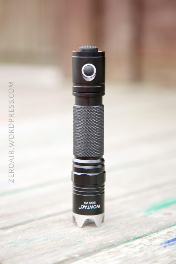 A black Wowtac BSS V3 flashlight stands upright on a wooden surface, with a textured grip and metal bezel, photographed in natural light.