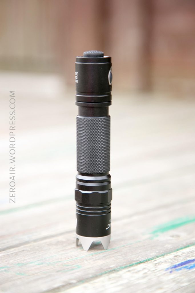 A black tactical flashlight with a textured grip stands upright on a wooden surface. The background is blurred, and ZEROAIR.WORDPRESS.COM is written vertically along the left side of the image.