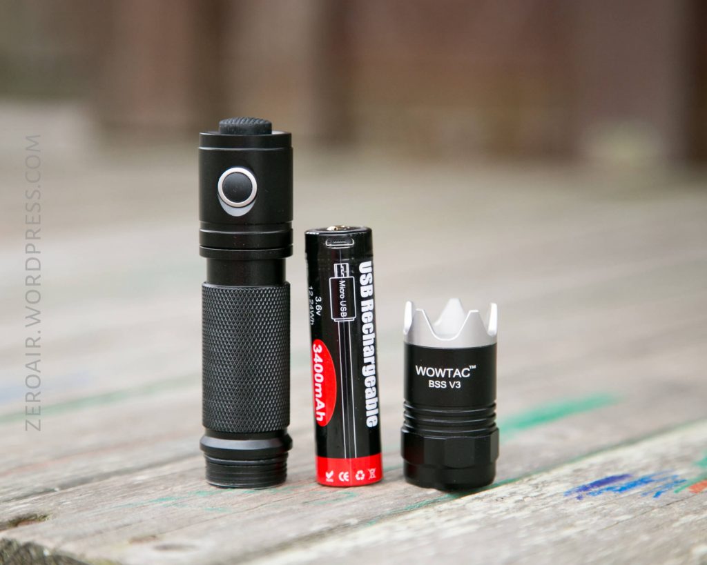 A disassembled black flashlight stands next to a red 18650 rechargeable battery and a silver-bezeled flashlight head marked WOWTAC BSS V3 on a wooden surface.