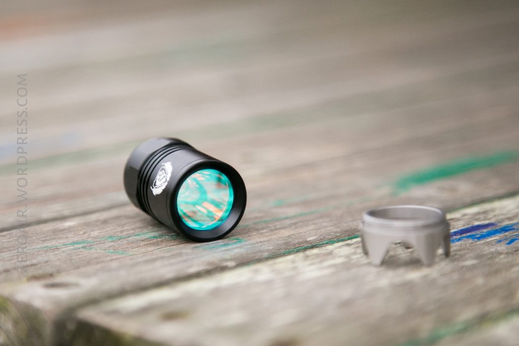 A small black flashlight with a reflective lens lies on a weathered wooden surface next to a detached silver metal bezel.