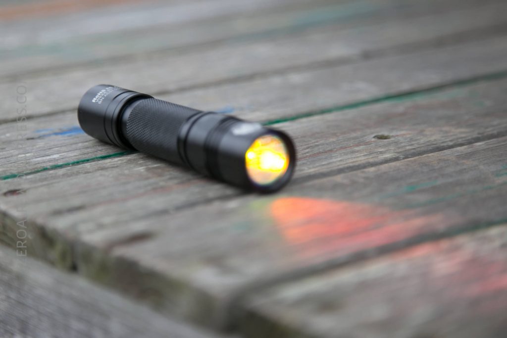 A small black flashlight lies on a weathered wooden surface, turned on and emitting a yellowish light that creates a faint glow and reflection on the wood.