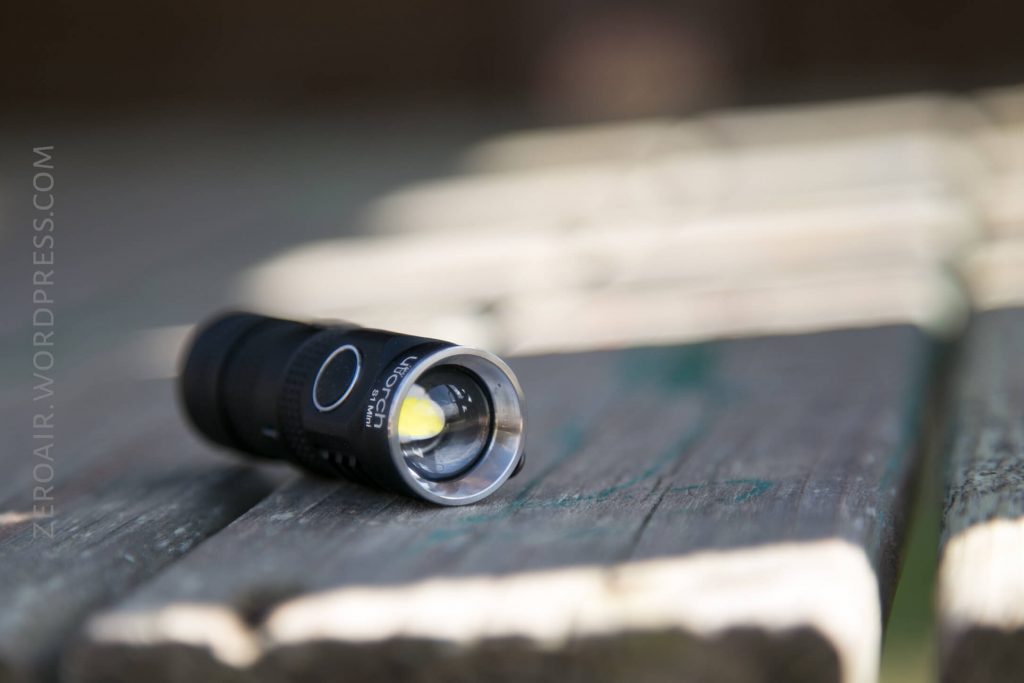 A small black flashlight with a silver rim around the lens lies on a weathered wooden surface next to an out-of-focus slatted wooden bench. The flashlight is positioned horizontally.