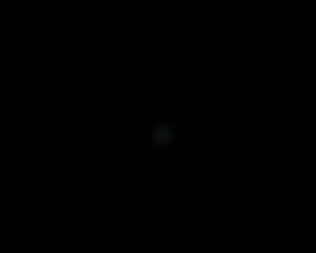 A mostly black image with a very faint, small, blurry gray-green dot near the center. No other visible details or objects.