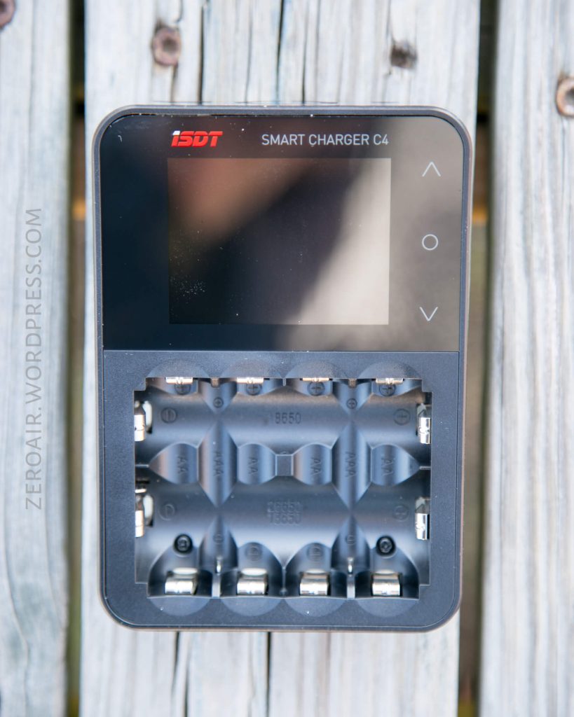 A close-up of an ISDT Smart Charger C4, showing its screen, control buttons, and four battery slots, placed on a wooden surface. The text ZEROAIR.WORDPRESS.COM is visible along the left side.