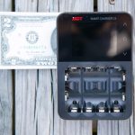 A two-dollar bill lies next to an ISDT Smart Charger C4, which has an empty battery compartment and a blank display screen, on a wooden surface.