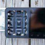 A two-dollar bill is placed under a black battery charger with four battery slots and a digital display, sitting on a weathered wooden surface.