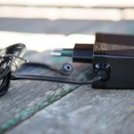 A black power adapter with a connected cable is placed on a wooden surface. The plug and barrel connector are visible, and the wire is loosely coiled beside the adapter.