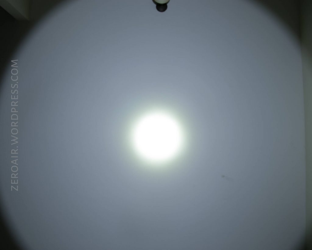 A bright white flashlight beam shines on a plain gray wall, creating a circular pattern with a bright center and fading edges. The words ZEROAIR.WORDPRESS.COM are visible along the left side of the image.