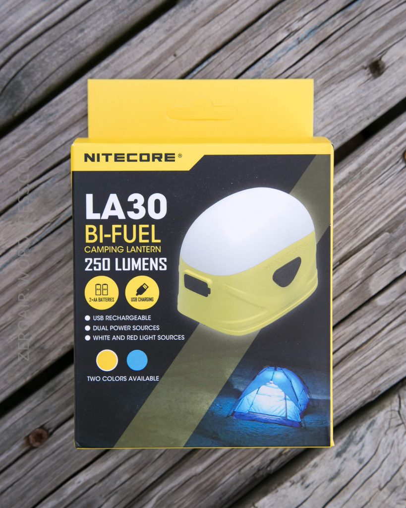 A Nitecore LA30 Bi-Fuel camping lantern box on wooden planks. The packaging highlights features such as 250 lumens, USB rechargeability, dual power sources, white and red lights, and two color options.