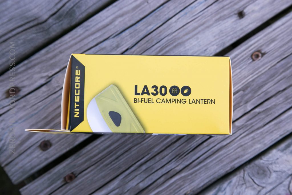 A yellow box labeled Nitecore LA30 BI-FUEL CAMPING LANTERN sits on a wooden surface. The box displays an image of the lantern and partial product details.