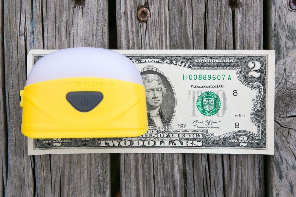 A small yellow Nitecore lantern sits on top of a two-dollar bill, both placed on a weathered wooden surface.
