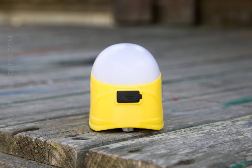 A small, yellow, dome-shaped portable lamp with a white top sits on a wooden surface. The lamp has a visible charging port covered by a black rubber flap on the front.