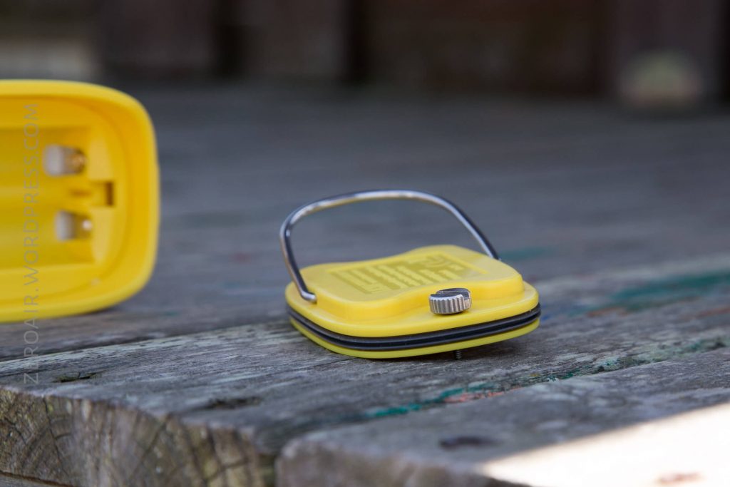 A small yellow device with a metal handle and a silver knob rests on a wooden surface. Part of another yellow object with prongs is visible on the left side of the image.