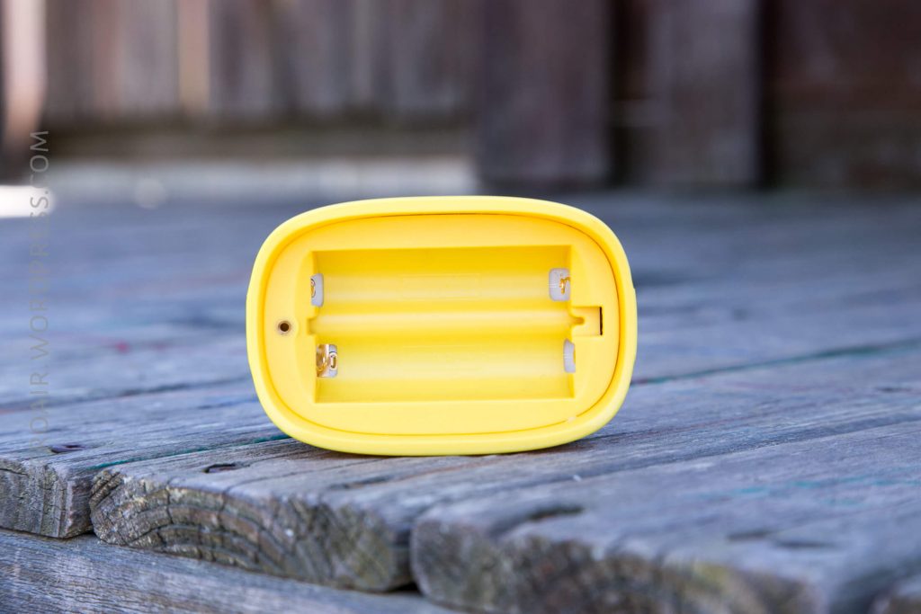 A yellow plastic battery compartment with two empty slots, placed on a weathered wooden surface outdoors.