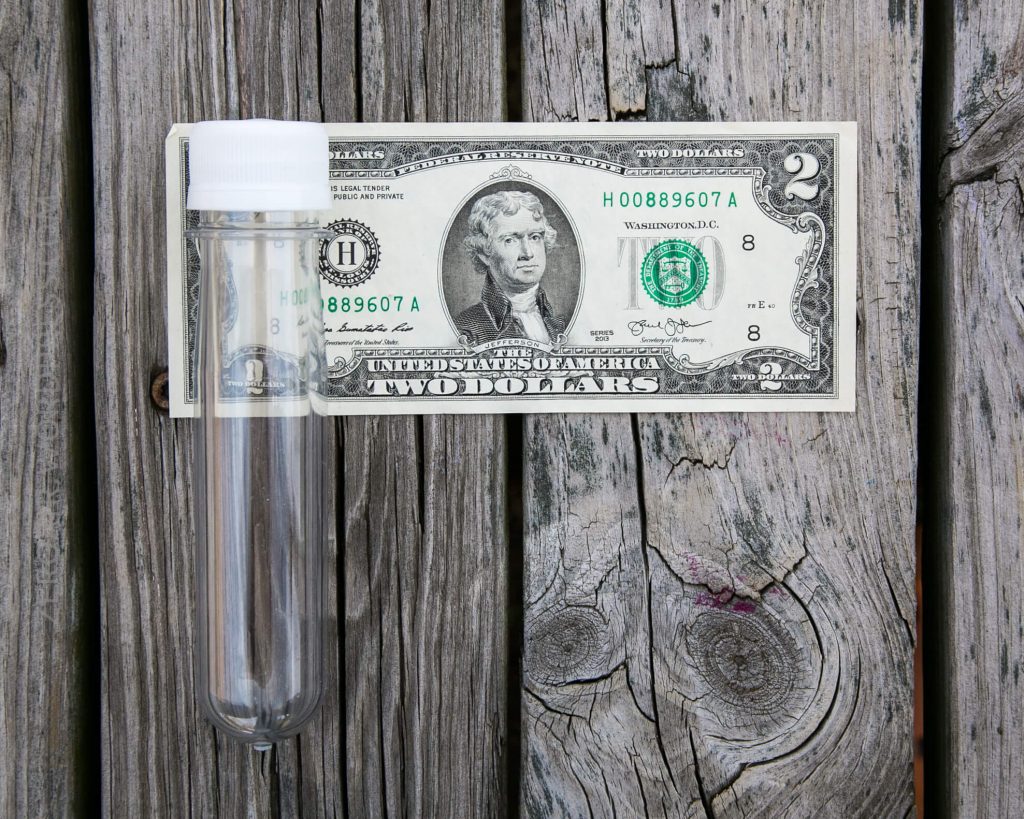 A clear plastic test tube with a white cap is placed next to a U.S. two-dollar bill on a weathered wooden surface. The bill features a portrait of Thomas Jefferson.
