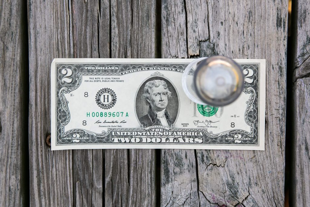 A two-dollar bill placed on weathered wooden boards with a clear glass knob partially covering the right side of the bill.