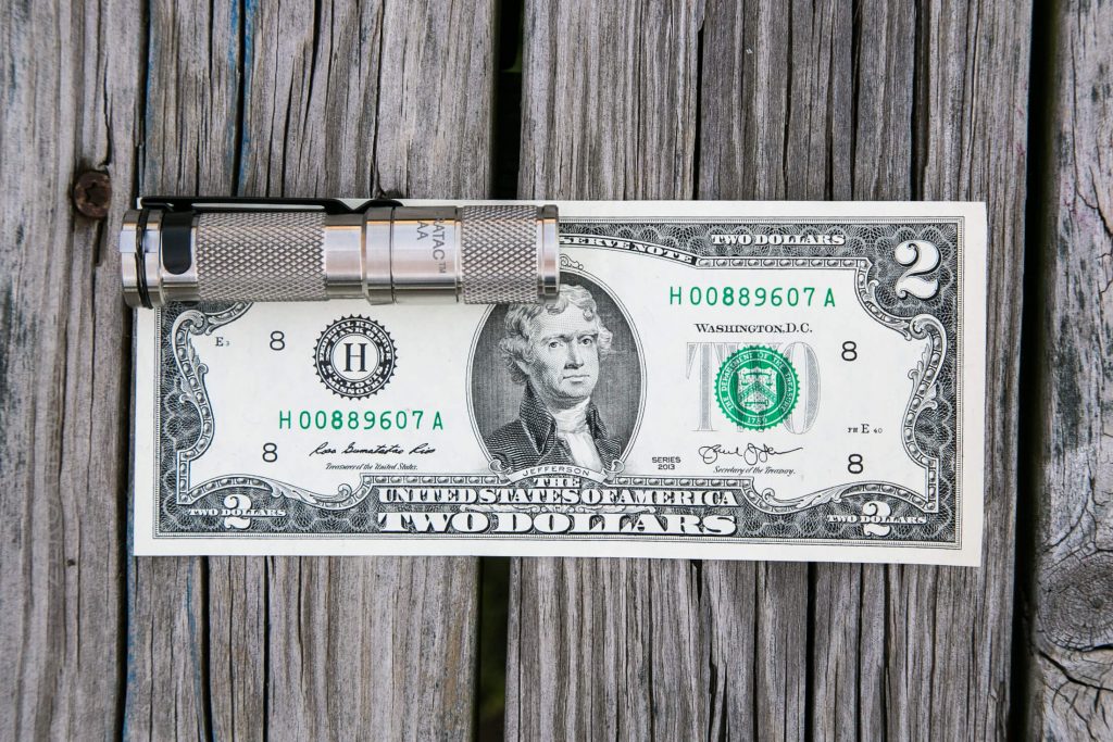 A silver flashlight is placed on top of a United States two-dollar bill, both resting on a weathered wooden surface.
