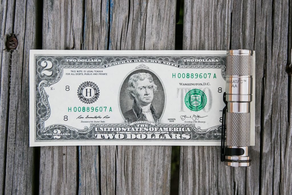 A US two-dollar bill lies flat on a wooden surface next to a silver flashlight, with both items positioned horizontally and parallel to each other.
