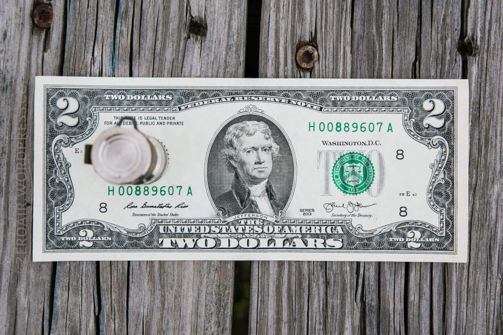 A US two-dollar bill with a bottle cap placed on its left side, lying on a weathered wooden surface with vertical gaps between the planks.