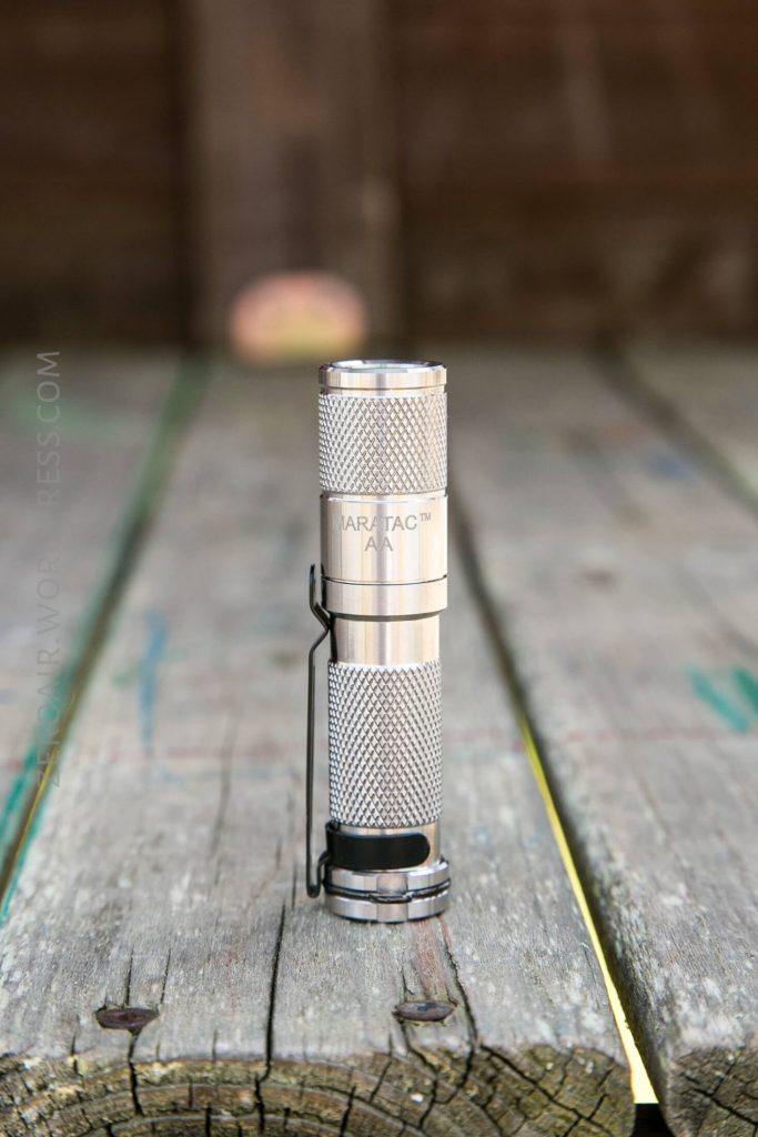 A small, silver Maratac AA flashlight stands upright on a weathered wooden surface, with a blurred brown background. The flashlight has a textured grip and a black pocket clip.