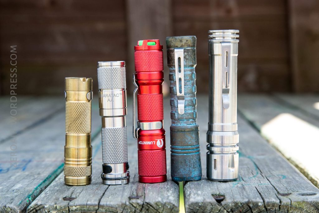 Five compact flashlights in various finishes—brass, silver, red, weathered blue, and stainless steel—stand upright on a worn wooden surface with a blurred background.