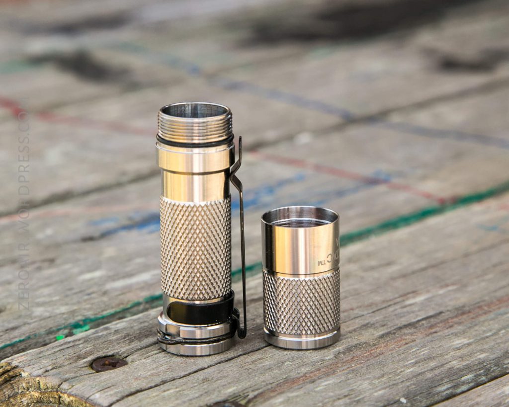 A small, silver metal flashlight stands upright on a wooden surface with its battery compartment open and the bottom cap placed beside it. The flashlight has a textured grip and a pocket clip.