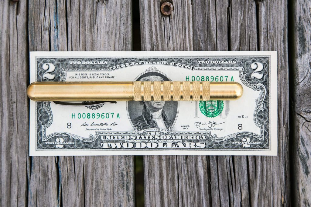 A two-dollar bill is placed on a weathered wooden surface with a gold-colored metal pen resting horizontally across its center.