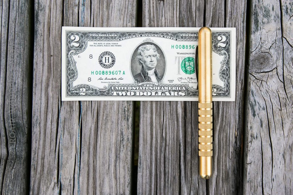 A two-dollar bill and a gold-colored pen are placed side by side on a weathered wooden surface, with the bill lying flat and the pen positioned vertically beside it.