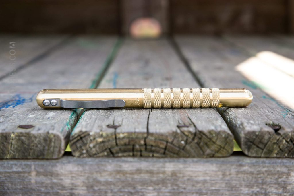 A brass pen with a textured grip and metal clip rests horizontally on a weathered wooden surface with visible grain and cracks. The background is softly blurred.