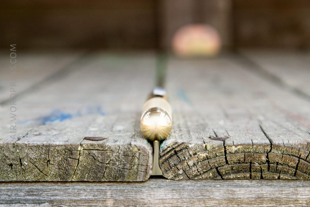 A close-up photo of a clear pen lying horizontally on the seam between two weathered wooden planks, with the background out of focus.