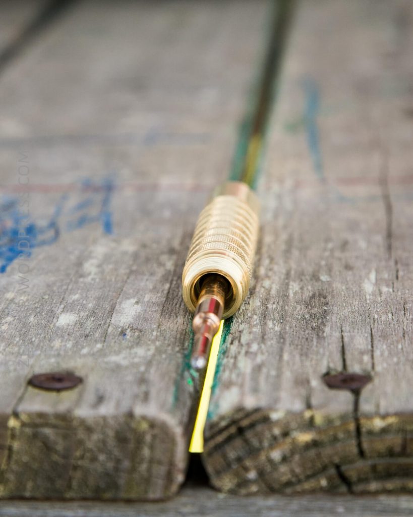 Close-up of a fishing rod with a cork handle resting on a weathered wooden surface, positioned so the rod points away from the camera along a gap in the wood.