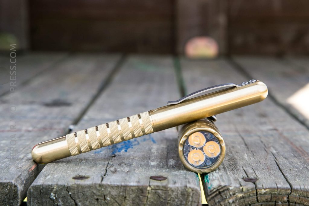 A brass flashlight with a textured grip and clip is balanced horizontally on top of a wooden surface, with its lens end showing three LED emitters arranged in a triangular pattern.