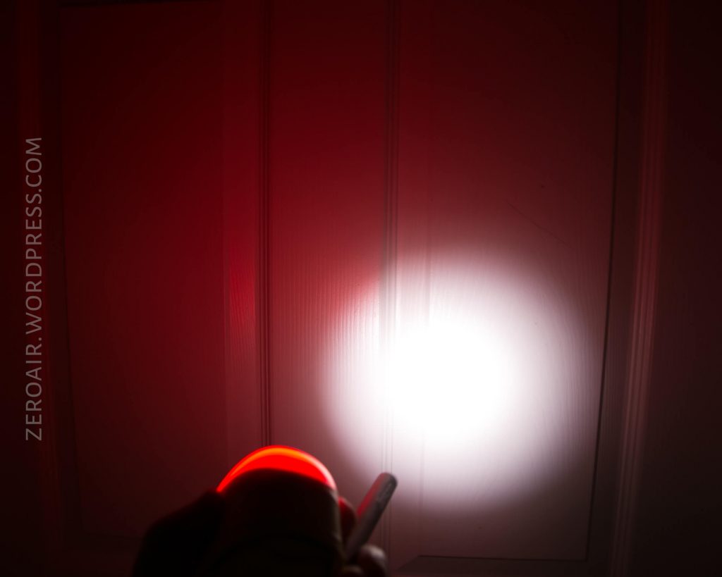 A flashlight with a red glowing ring projects a bright white beam onto a white paneled door in a dimly lit environment. The text ZEROAIR.WORDPRESS.COM appears along the left side.