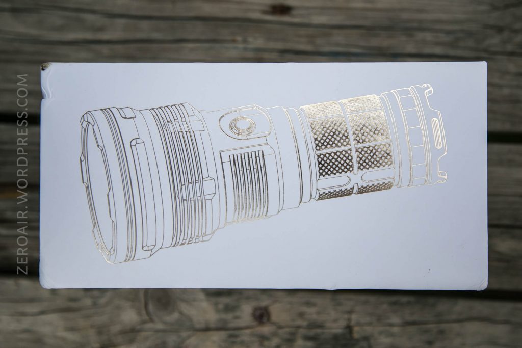 A white box with a gold line drawing of a large, cylindrical flashlight on the top. The background is wooden planks. The text “ZEROAIR.WORDPRESS.COM” is printed vertically along the left side.