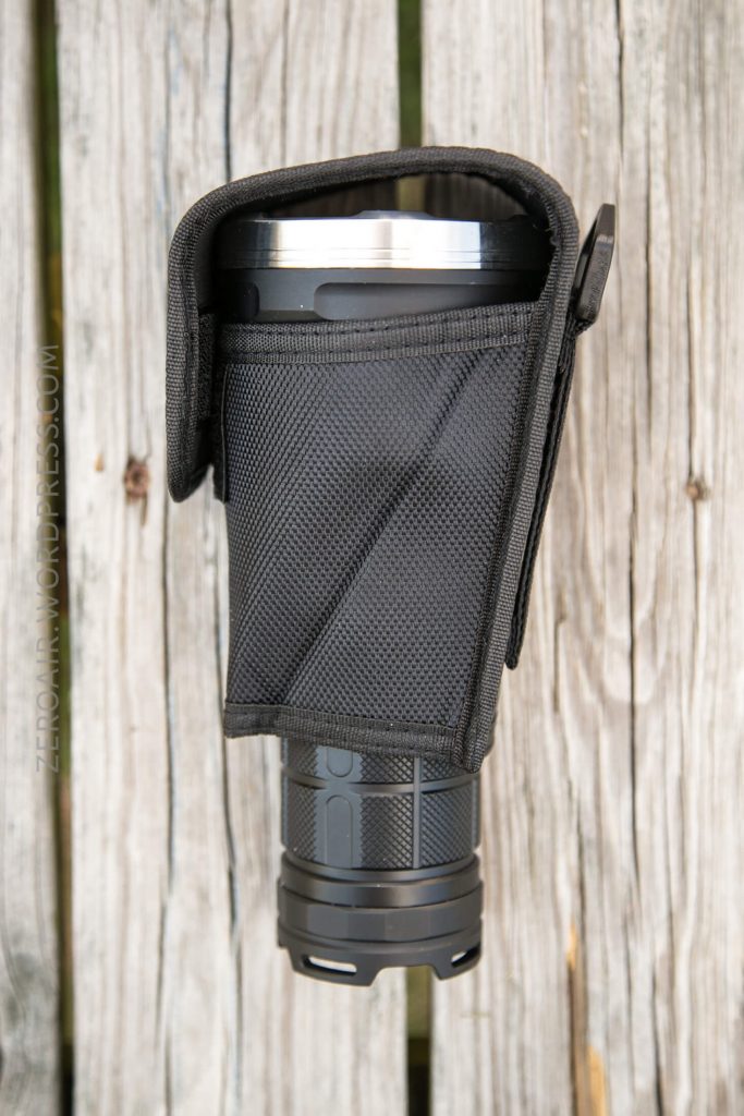 A large flashlight partially inserted in a black nylon holster with a flap, resting vertically on weathered wooden boards.