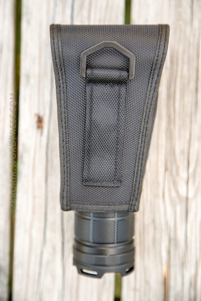 A black, textured nylon pouch with a stitched belt loop and metal clip is shown vertically on a wooden surface. The bottom of a cylindrical black object is partially visible inside the pouch.