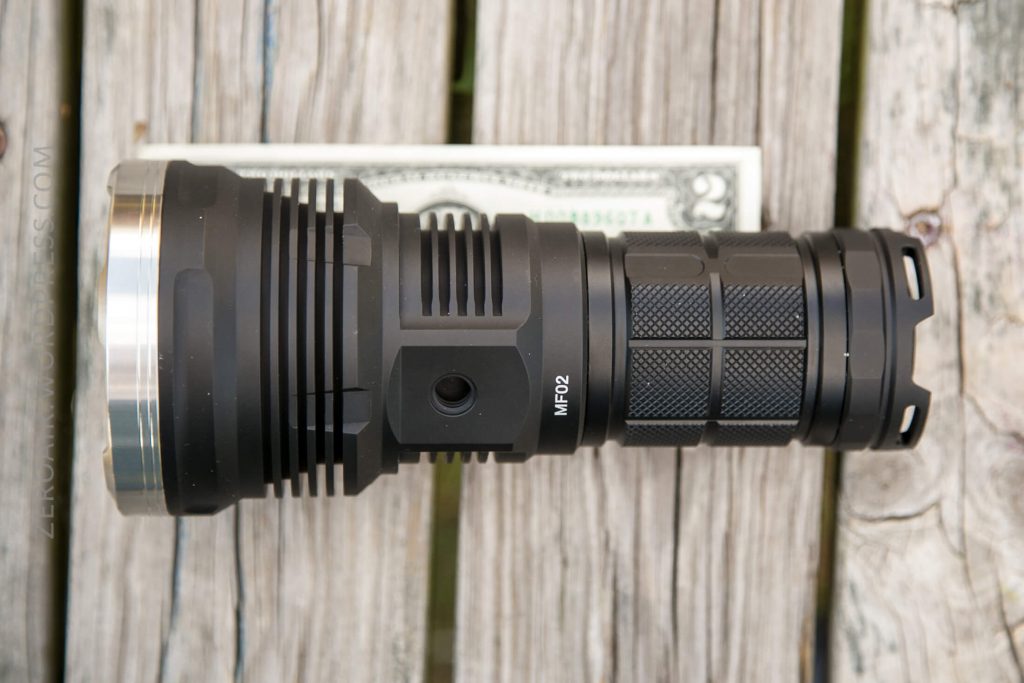 A black tactical flashlight labeled MF02 lies on a weathered wooden surface, with a U.S. dollar bill partially visible underneath for size comparison.