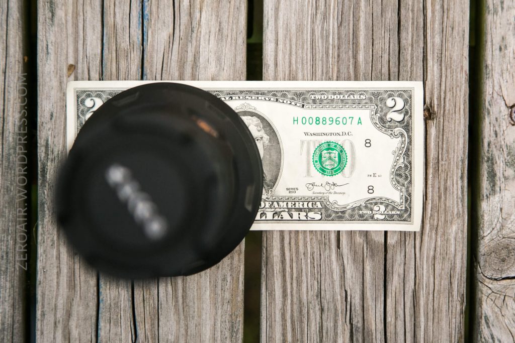 A two-dollar bill is placed on a weathered wooden surface, partially covered by a camera lens cap in the foreground. The bill’s serial number and details are clearly visible.