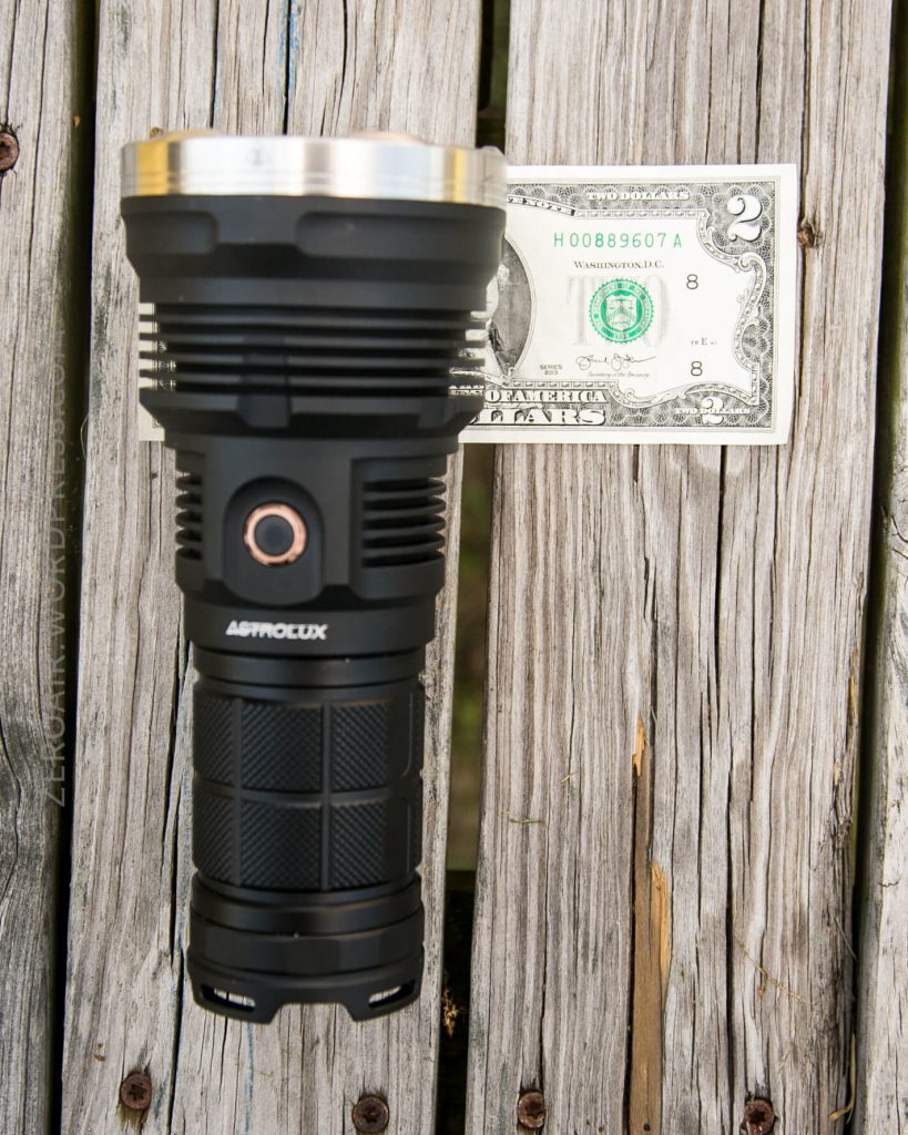 A black Astrolux flashlight is placed vertically next to a two-dollar bill on a weathered wooden surface, showing the flashlight’s size in comparison to the bill.