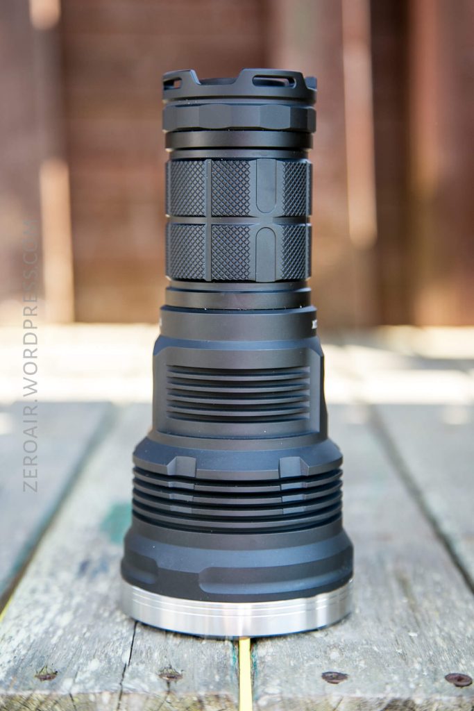 A large, black, heavy-duty flashlight with a textured grip is standing upright on a weathered wooden surface. The background is blurred, showing wooden planks and outdoor lighting.