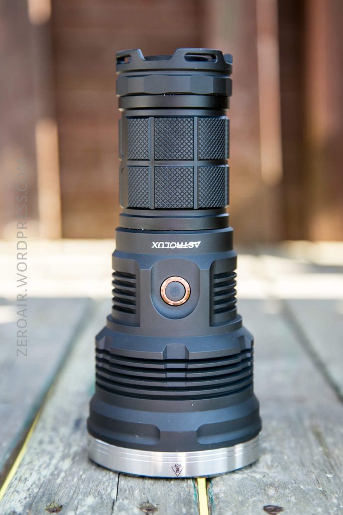 A black Astrolux flashlight stands upright on a wooden surface outdoors, featuring a textured grip, copper-colored button, and silver base with engraved details.
