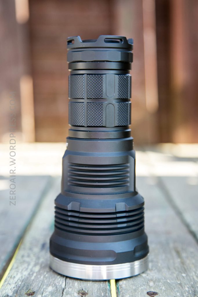 A large, black flashlight with a textured grip and metal base is standing upright on a wooden surface. The background is out of focus, showing wooden boards.
