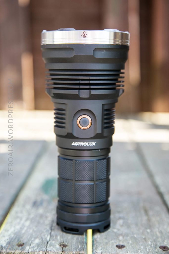 A black Astrolux flashlight stands upright on a weathered wooden surface outdoors, with a blurred brown background.