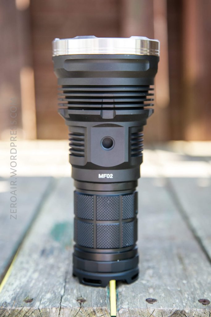A large black flashlight labeled “MF02” stands upright on a weathered wooden surface with a blurred brown and green background.